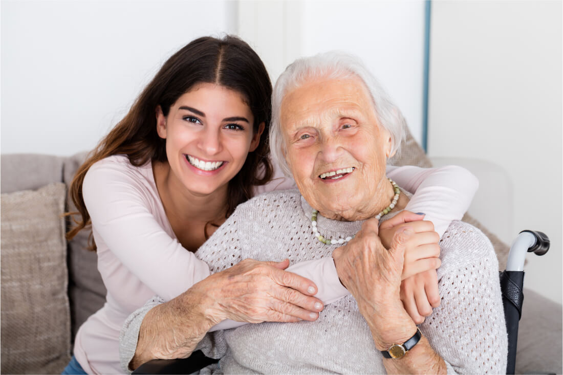 Elderly lady sitting in a wheelchair smiling with her smiling caregiver hugging her