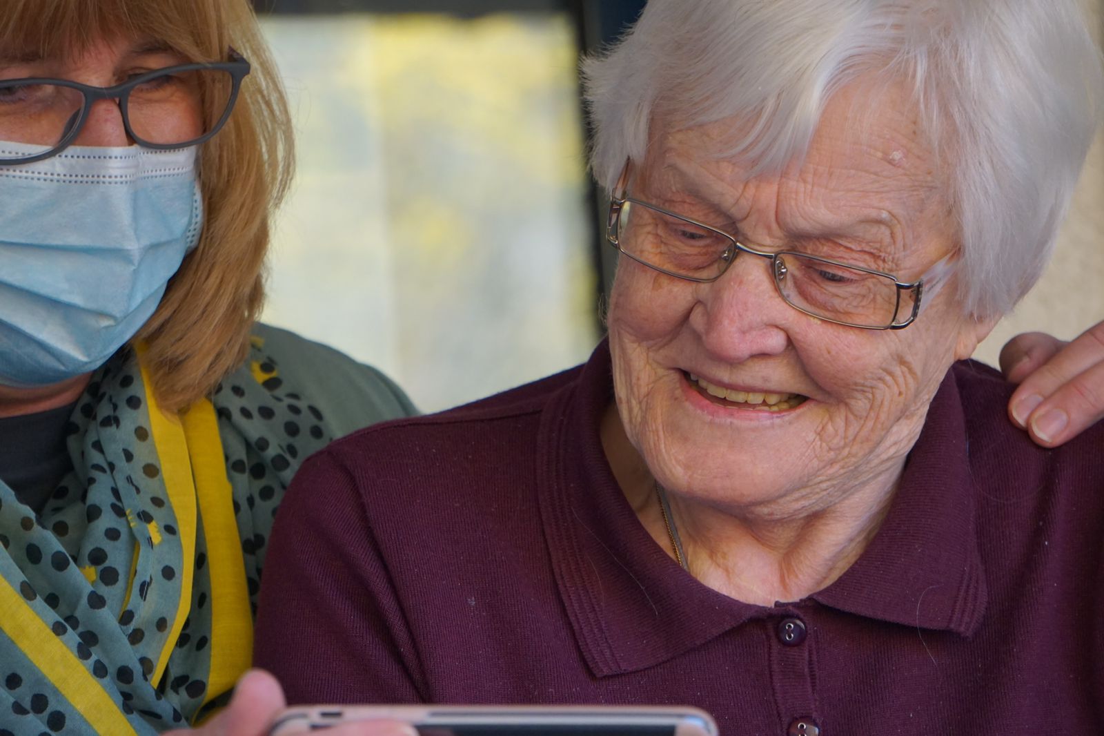 A caregiver wearing a mask is sharing something on her phone to an elderly lady smiling